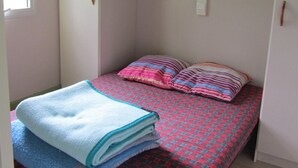 A small, single-bed room with a red and blue checkered bedspread, a blue blanket, and two striped pillows. There is a window on the left, a white wardrobe, and a small white shelf unit above the bed. - Camping des Etoiles - Chalets (Najac)