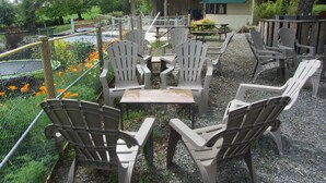 Outdoor seating with Adirondack chairs and a small table, surrounded by a gravel area and a fence. - Camping des Etoiles - Chalets (Najac)