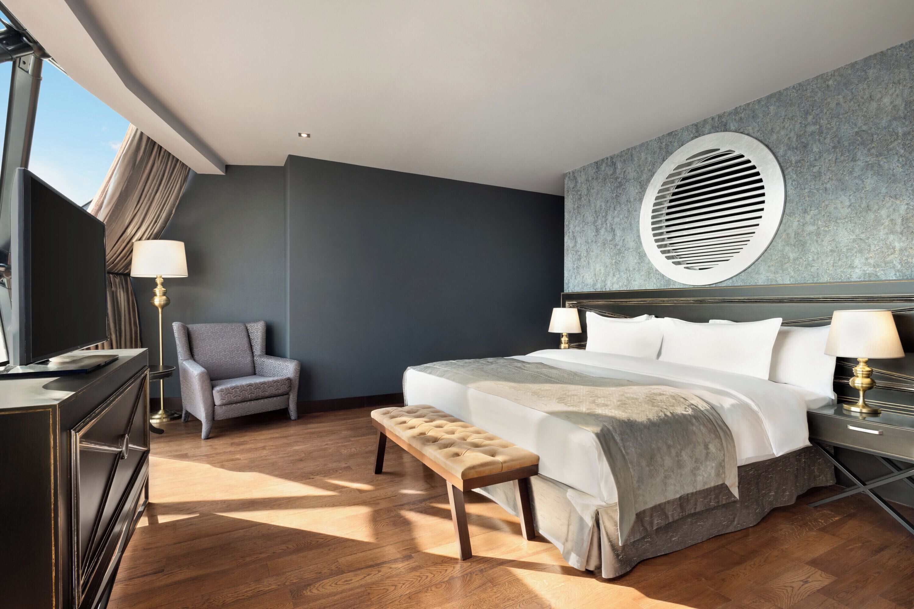 A hotel room with a large bed, a bench, a chair, a desk, and a window with curtains.
