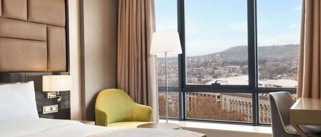 Une chambre d’hôtel avec un grand lit, un bureau, une chaise et une vue sur le paysage urbain, visible à travers une grande fenêtre.