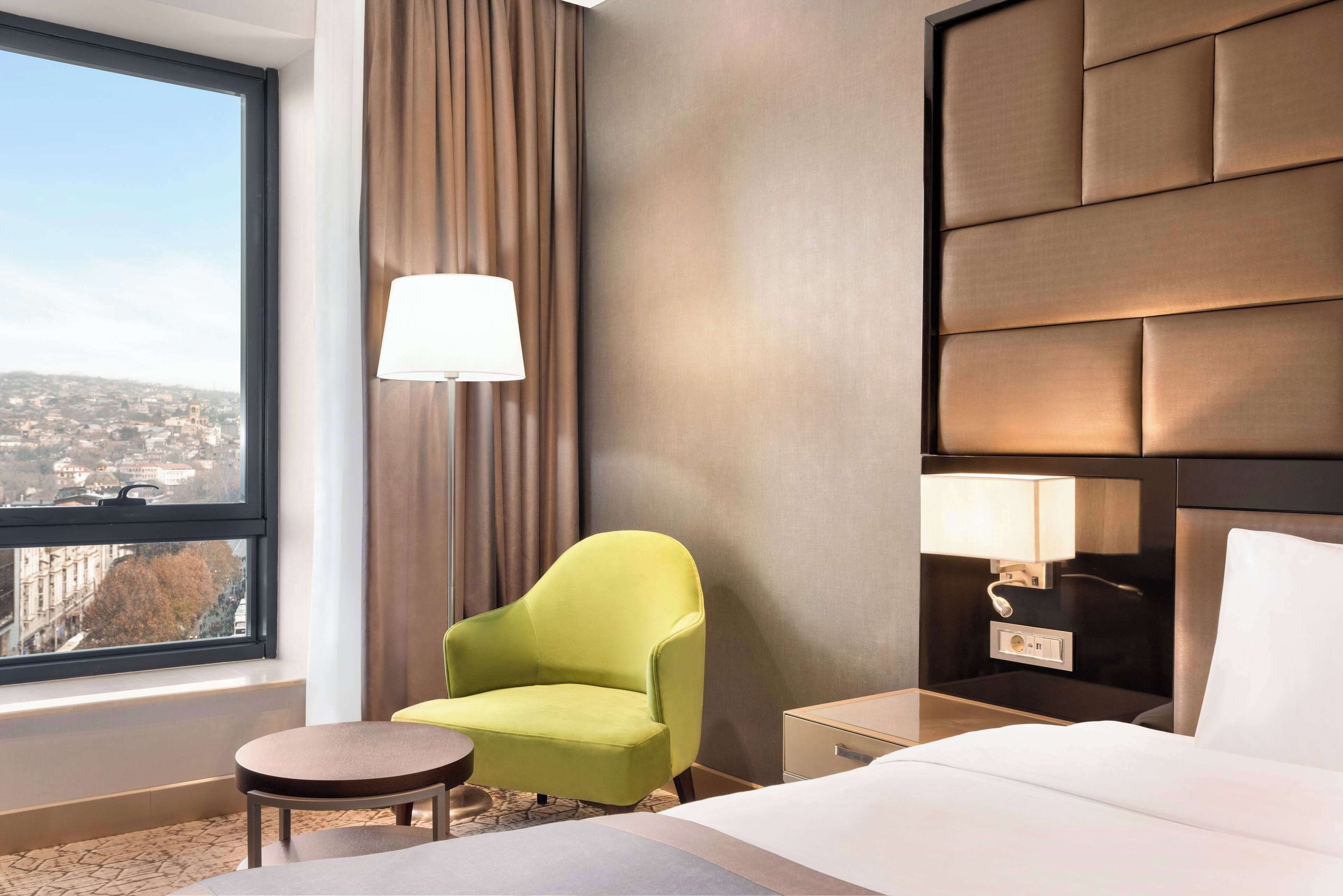 A hotel room with a large window, a green armchair, a bedside table with a lamp, and a bed with a headboard.