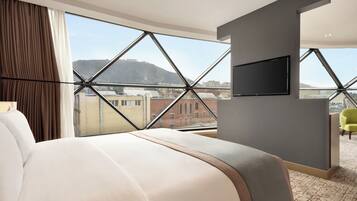 Habitación de hotel con una cama grande, un televisor montado en la pared y vistas a edificios y colinas a través de amplios ventanales.