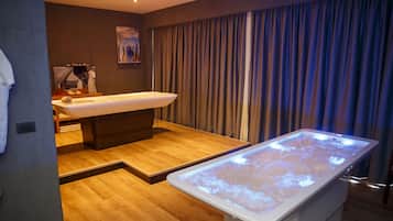 A modern spa room with a massage table, a large bathtub, and a wooden floor.
