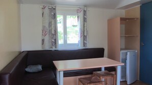 A compact living space with a sofa, a small table, two chairs, a refrigerator, and a window with curtains. - Camping des Etoiles - Mobil-Home (Najac)