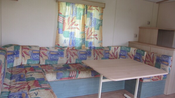 A compact living space with a sofa and a small table, a window with curtains, and a kitchenette area. - Camping des Etoiles - Mobil-Home (Najac)
