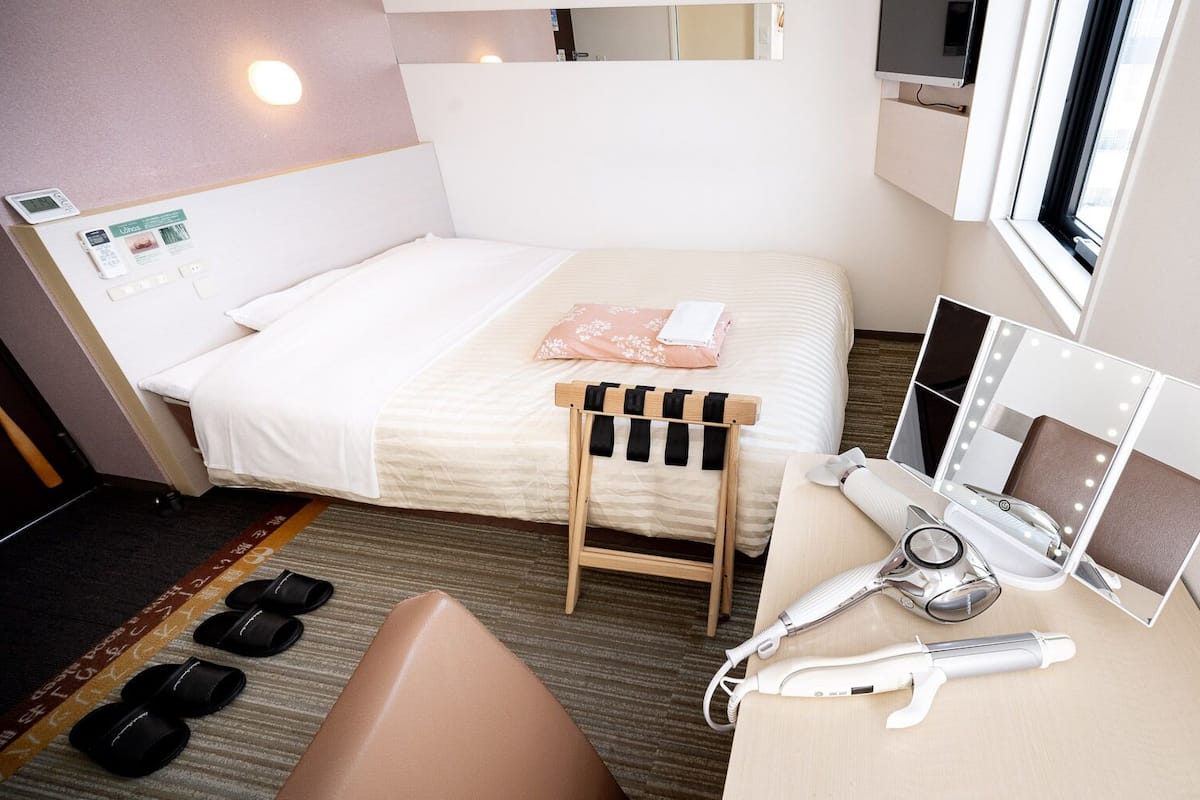 Une chambre d’hôtel comprenant un lit, une petite coiffeuse avec un miroir et des lampes, un sèche-cheveux et un porte-chaussures.