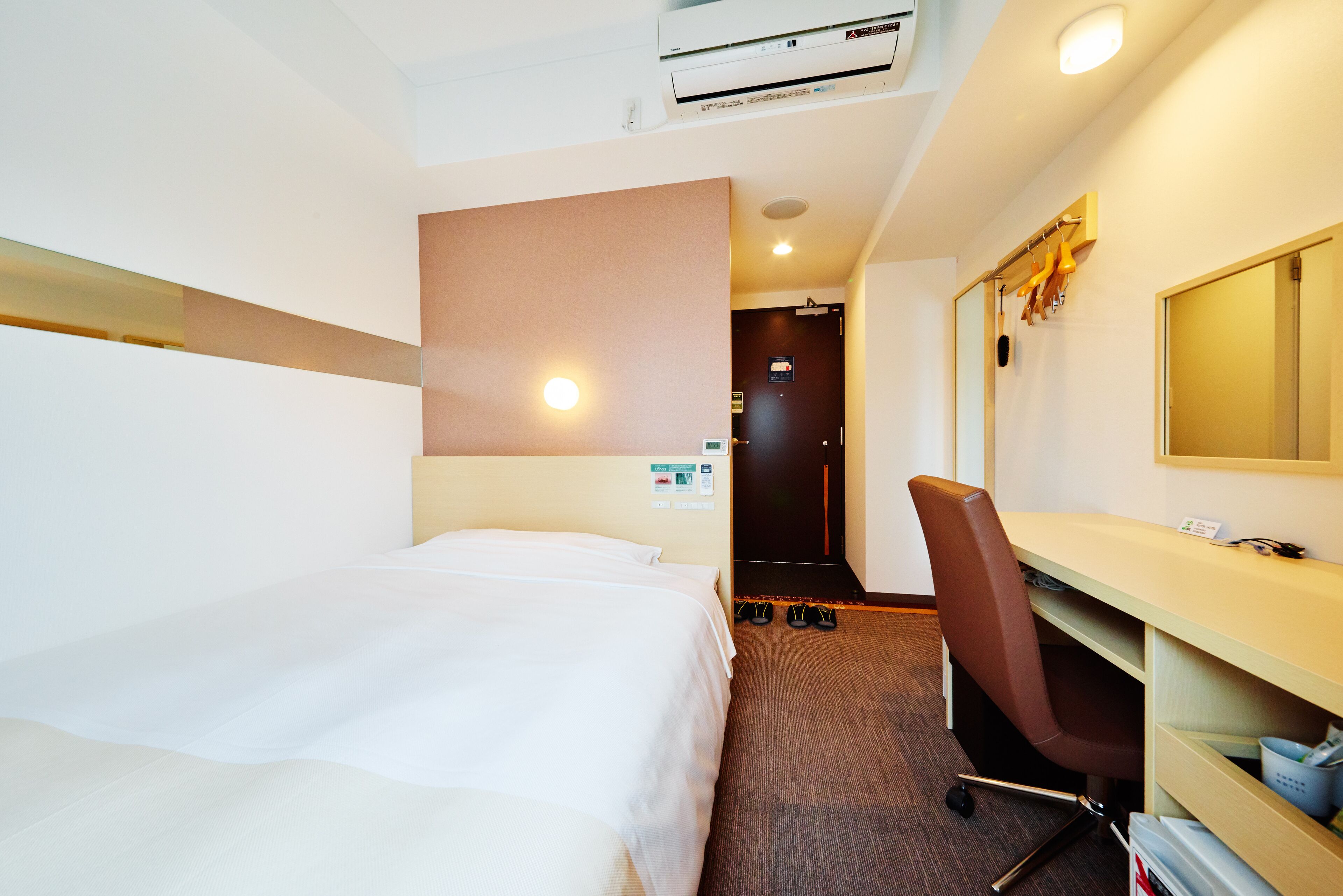 A hotel room with a bed, a desk, a chair, a mirror, and a shoe rack.