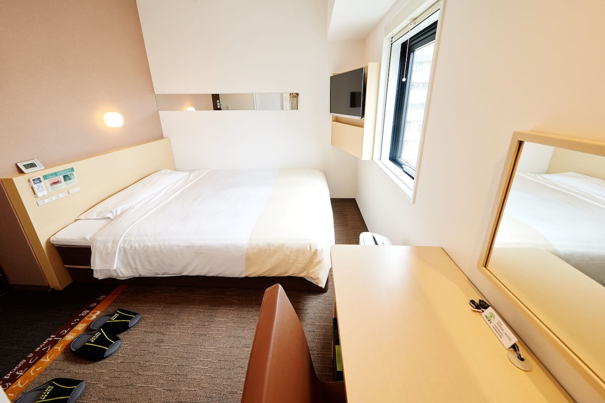 Une chambre d’hôtel équipée d’un lit, d’un bureau, d’une chaise, de chaussons, d’une télévision et d’une fenêtre.