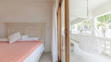 A bedroom with a bed, a balcony with a white chair, and a view of palm trees.