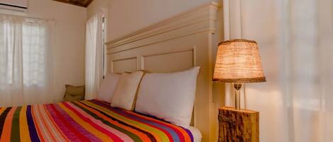 Una cama con un edredón de rayas de colores, una mesita de noche de madera con una lámpara y un cabecero blanco.