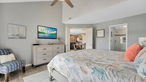 Une chambre à coucher comprenant un lit, une commode, une chaise et un bureau avec une autre chaise. Il y a un tableau accroché au mur et un téléviseur placé au-dessus de la commode.