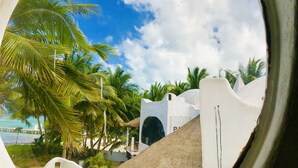 A view through a circular window of a coastal area with palm trees, a white building, and a pier in the background. - Beautiful Suite double beach front (Cancun)