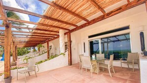 Un patio techado con un pergolado de madera, mobiliario exterior y vista al océano.