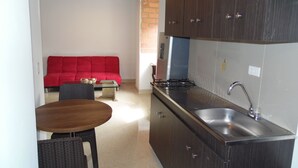 A compact kitchen with dark wooden cabinets, a stainless steel sink, and a gas stove. There's a red sofa and a small dining table with chairs in the background. - Studio in Laureles (Medellín)