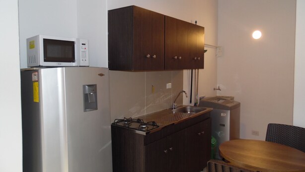 A compact kitchen with a refrigerator, microwave, stove, sink, and dark wooden cabinets. - Studio in Laureles (Medellín)