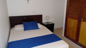A bed with a blue and white bedspread, a bedside lamp, and a wooden wardrobe with a cross on the wall. - Studio in Laureles (Medellín)