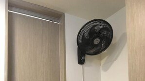 A room with a wooden door, a desk with a black chair, and a standing fan.