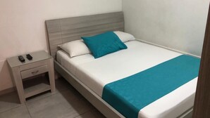 A neatly made bed with a teal throw blanket and pillows, a small bedside table with a drawer, and a remote control on the table.