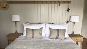 Cama bem arrumada com lençóis brancos e duas almofadas bege, acompanhada por duas mesas de cabeceira com luminárias, em frente a uma parede com painéis verticais.