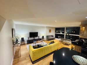 Una sala de estar moderna con un sofá amarillo, un televisor de pantalla plana y una zona de comedor con sillas y una mesa.