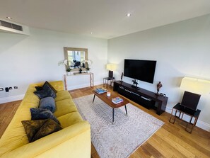 A living room with a yellow sofa, a coffee table, a TV, and a small side table. - Thames Point, Imperial Wharf (London)