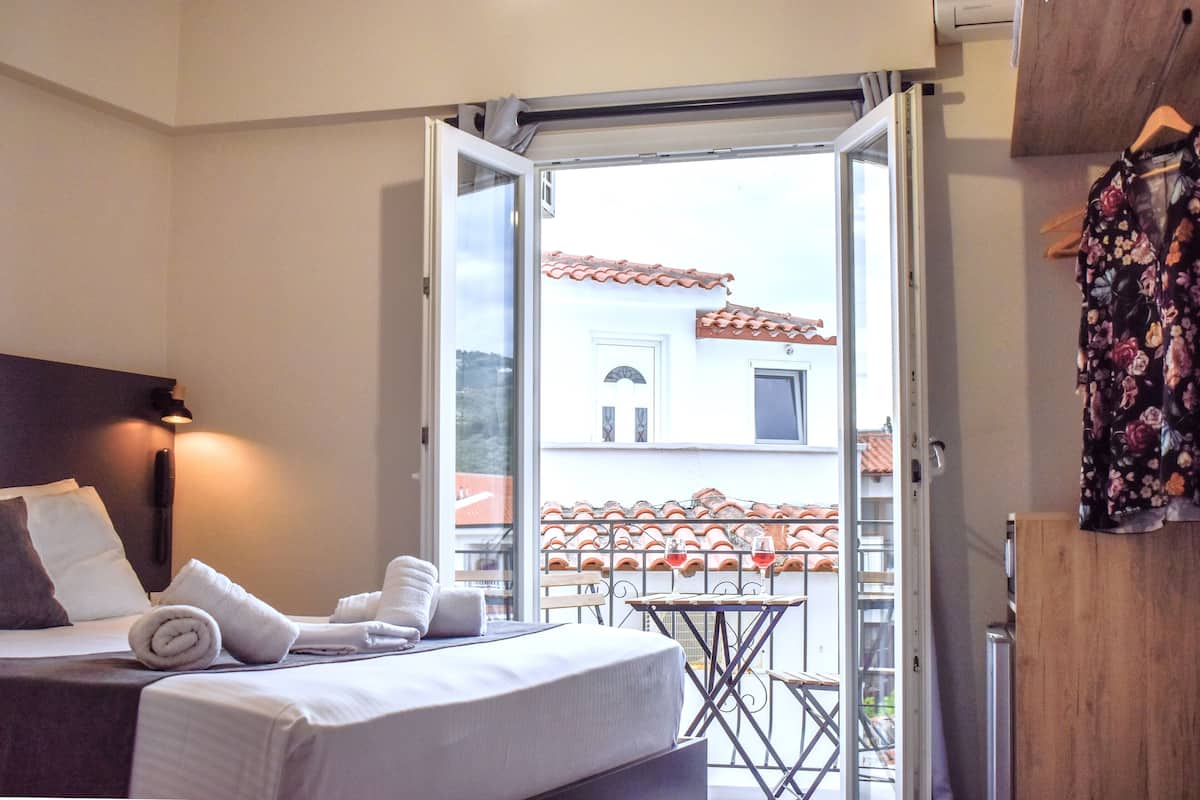A hotel room with a bed, a small table with two wine glasses, a balcony with a tiled railing, and a wooden door.