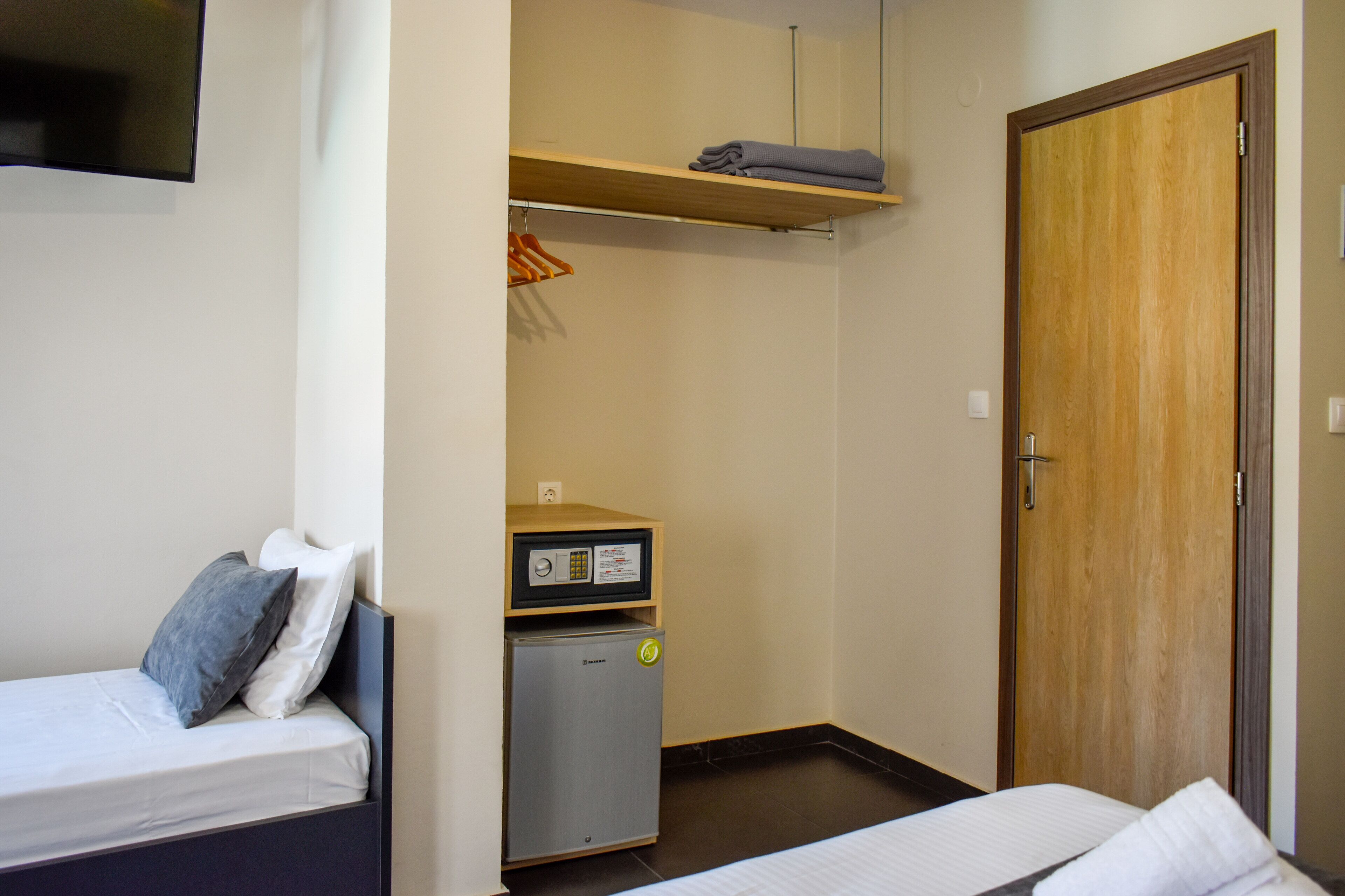 A compact hotel room with a bed, a small refrigerator, a wooden wardrobe, and a shelf with folded clothes.