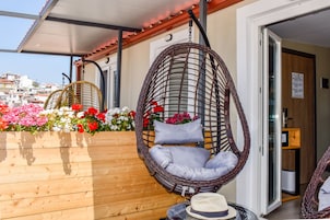 Una silla colgante de mimbre con cojines, una maceta de madera con flores y un sombrero sobre una mesa.