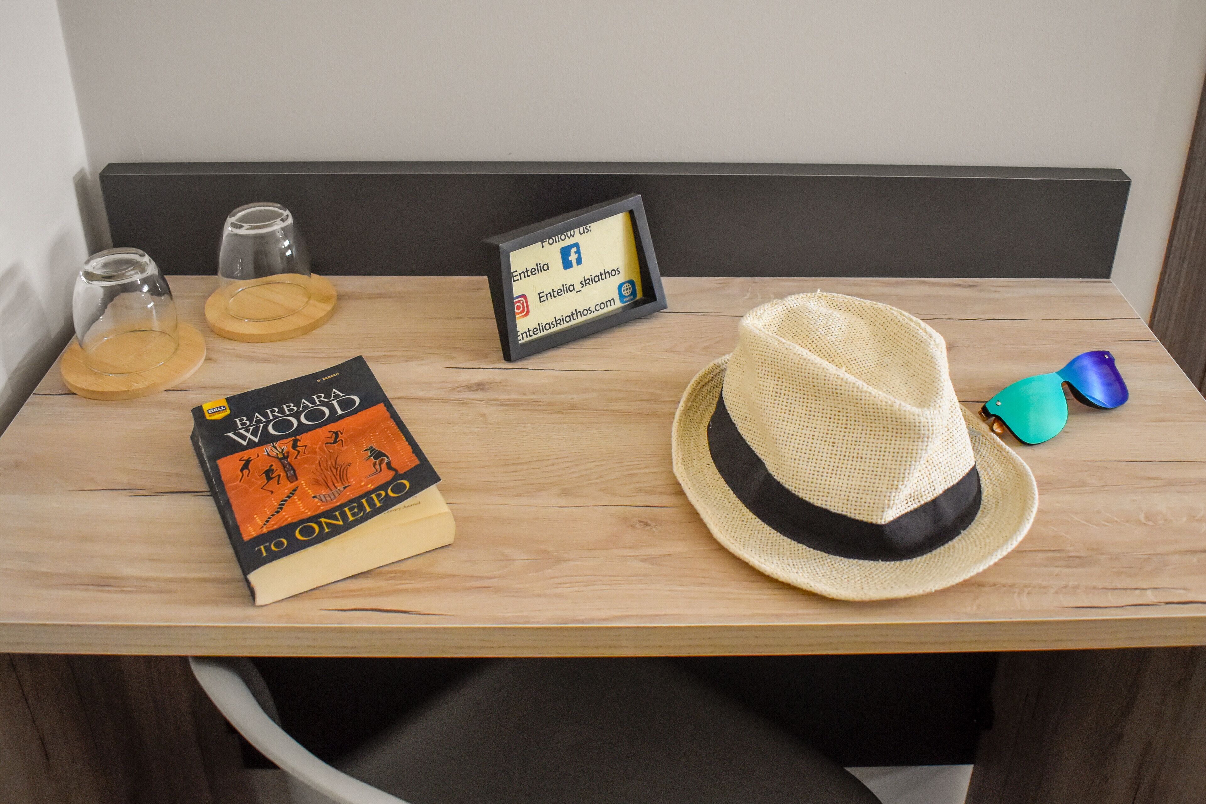 Een houten tafel met een boek getiteld 'To Oneiro', twee glazen, een lijst met een social media bericht en een strohoed.