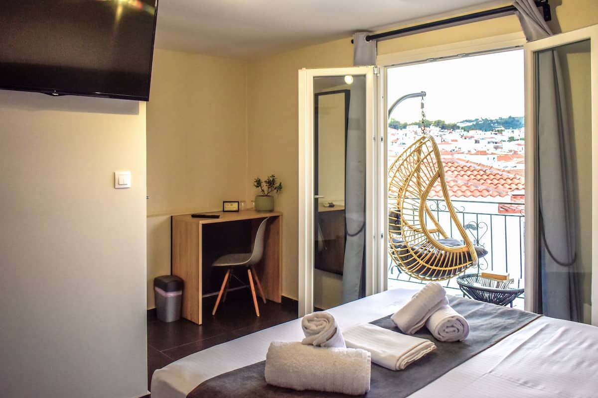 A hotel room with a bed, a desk, a chair, a small potted plant, and a balcony with a view of the city.