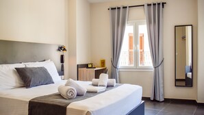 A neatly made bed with pillows and rolled towels, a bedside table with a lamp, a window with curtains, and a mirror on the wall.