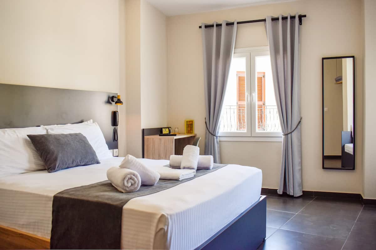 A neatly made bed with pillows and rolled towels, a bedside table with a lamp, a window with curtains, and a mirror on the wall.