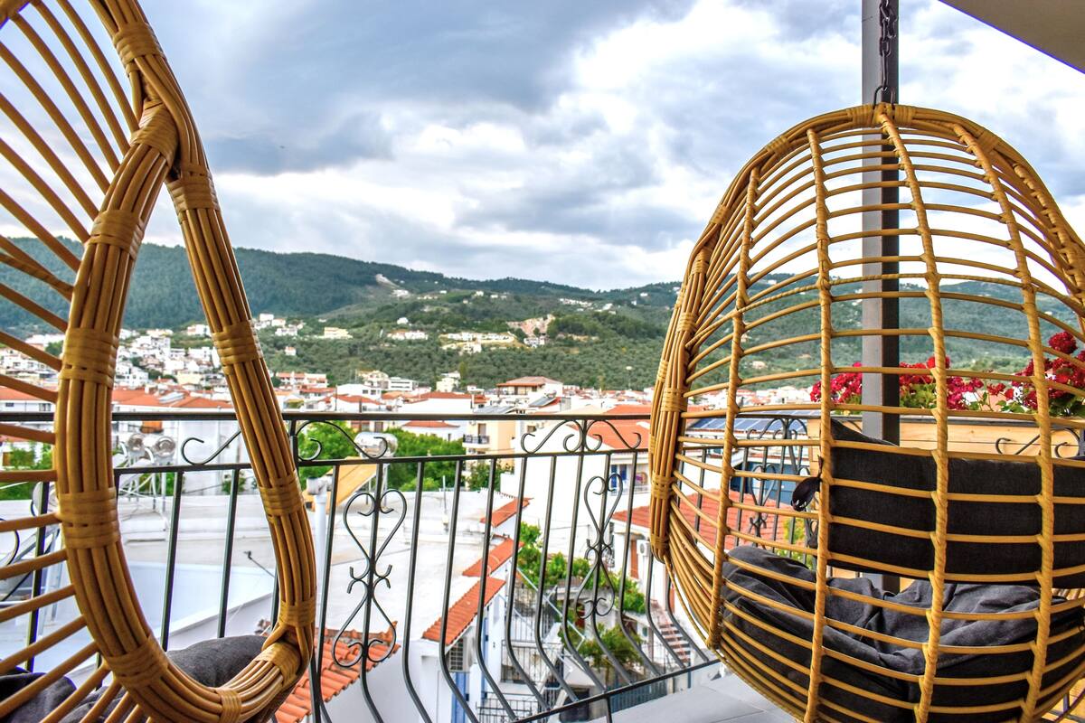 A wicker chair with a cushion on a balcony overlooking a town with red-roofed buildings and green hills in the distance.