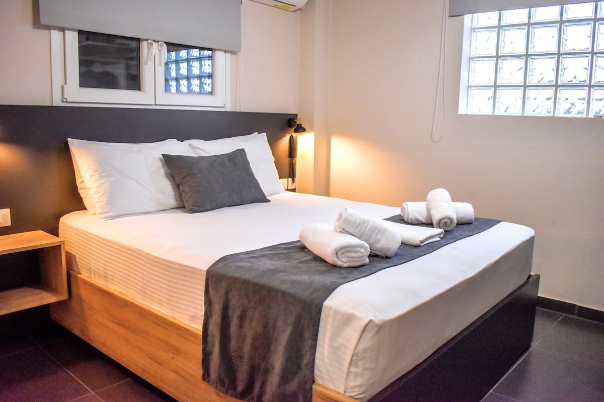 A neatly made bed with white and gray linens, a dark headboard, and a window with blinds.