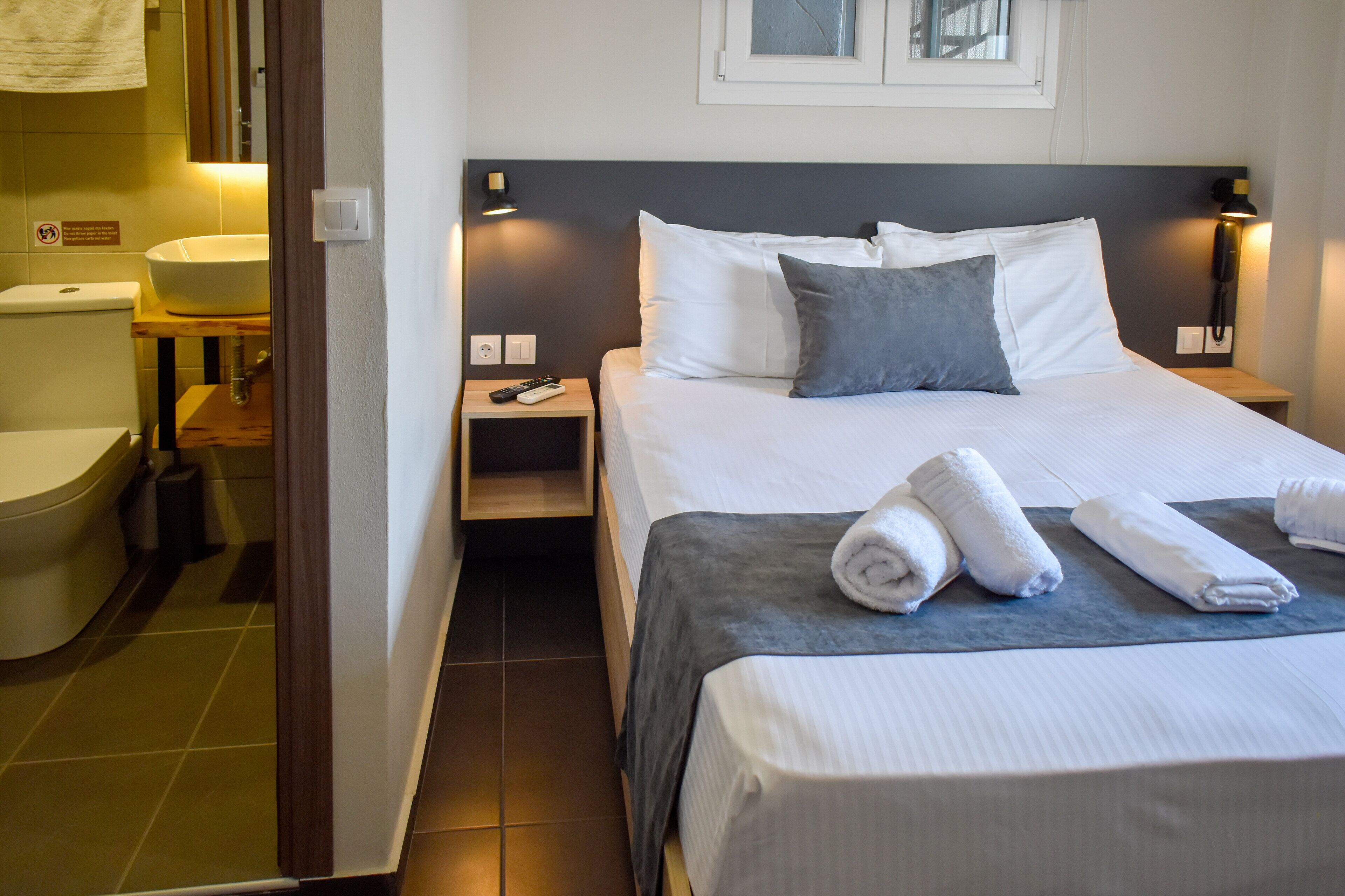 A hotel room with a bed, bedside tables, a nightstand, a bathroom with a toilet and sink, and a towel on the bed.