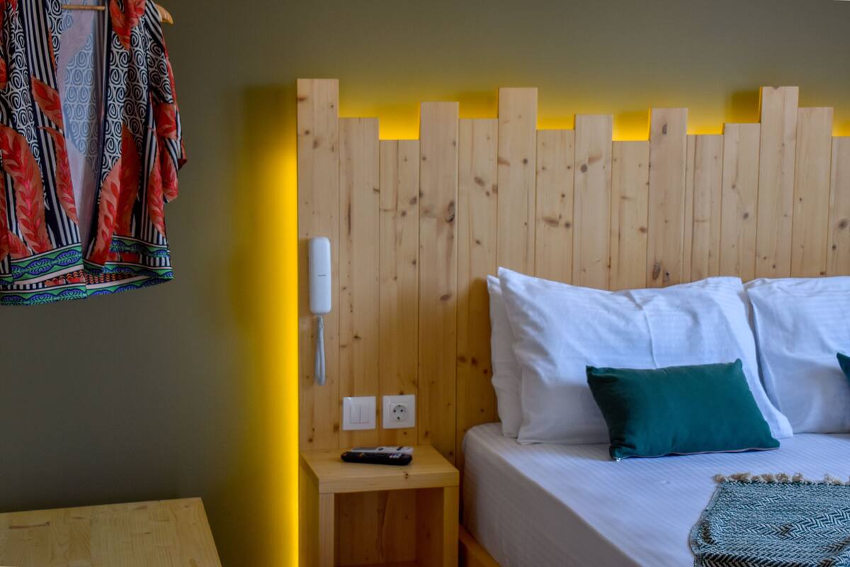A room with a wooden headboard, white bedding, a green pillow, a remote control on a wooden nightstand, and a telephone.