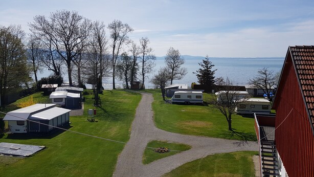 Ein Campingplatz mit Wohnmobilen und Wohnwagen am Seeufer, ein Kiesweg und Bäume.