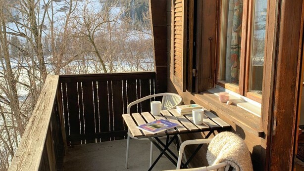 A wooden deck with a table and chairs, a window with curtains, and a view of trees and sky. - Apartment Iris by Interhome (Lenk)