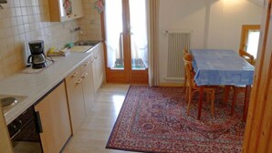 A kitchen with a coffee maker, a sink, and a window with curtains. There is a dining area with a table and chairs, and a patterned rug on the floor. - Heiderösli by Interhome (Grächen)