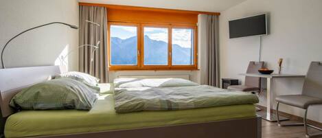 A hotel room with a bed, a desk, a chair, a TV, and a window with a mountain view.