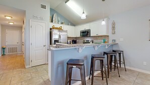 A kitchen with white cabinets, a stainless steel refrigerator, a microwave, and a granite countertop. There are three bar stools and a hanging pendant light. - FC1702 Private Heated  Pool, Room for Boat Parking, Golf Cart Included (Port Aransas)