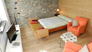 Une pièce chaleureuse avec un mur en pierre, un lit en bois, un fauteuil orange et une petite table avec une lampe.