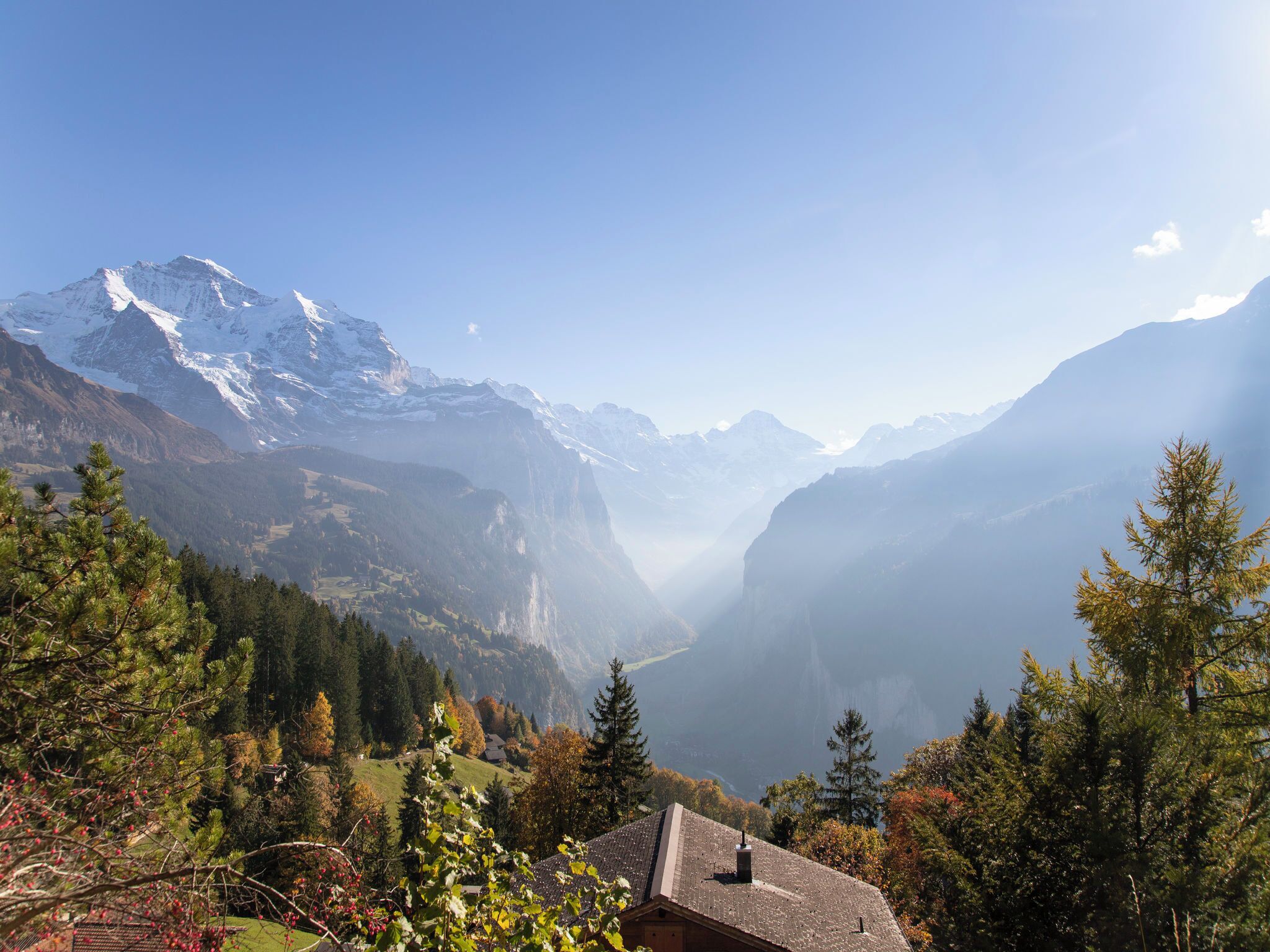 Eine Berglandschaft mit einem Haus im Vordergrund, umgeben von Bäumen und verschneiten Berggipfeln im Hintergrund.