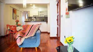 A modern living room with a blue sofa, a dining area with red chairs, and a kitchen in the background.