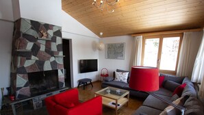 A living room with a stone fireplace, a flat-screen TV, a red armchair, a grey sofa, and a large window with curtains.