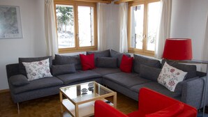 A living room with a grey sectional sofa, red cushions, a glass coffee table, and a red armchair. There are two windows with curtains, a red floor lamp, and a framed picture on the wall.