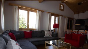 A living room with a grey sofa, red cushions, a glass coffee table, and a red armchair. There are windows with curtains, a red lamp, and a small table with a lamp.
