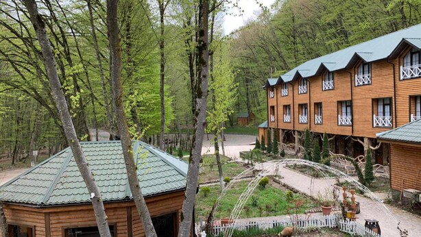 A wooden cabin with a green roof, surrounded by a garden with a white picket fence, and a larger building with balconies in the background. - Gachresh Forest Resort (Quba)