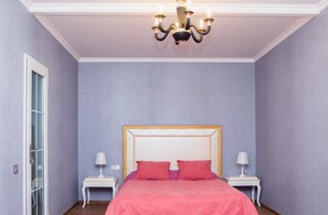 Un dormitorio con una cama grande, dos mesitas de noche con lámparas, una araña y una puerta con paneles de cristal.