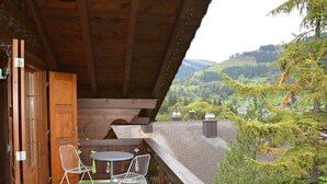 Un balcón de madera con dos sillas blancas y una mesita, con vistas a un valle con árboles y edificios.